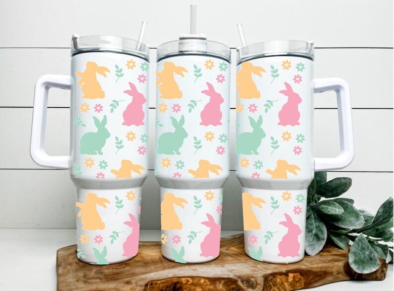 May include: Three white insulated tumblers with handles, each adorned with a repeating pattern of pastel-colored bunnies, flowers, and leaves. The tumblers have white lids and straws, perfect for cold beverages. The tumblers are approximately 10 inches tall.