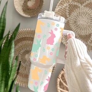 May include: White tumbler with a handle, decorated with pastel-colored bunny silhouettes, flowers, and leaves. The tumbler has a clear lid and a straw. The design includes yellow, pink, and mint green elements, perfect for Easter.