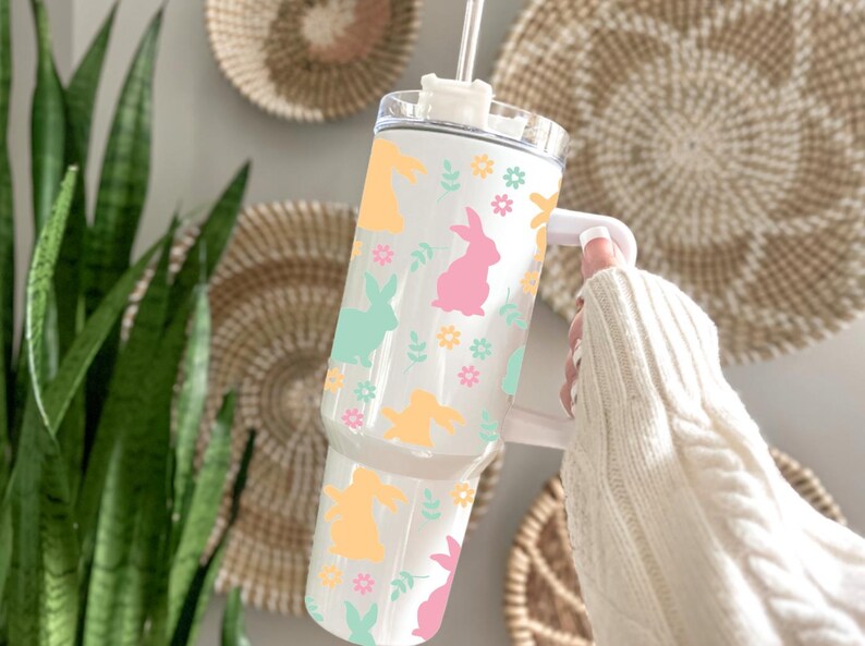 May include: White tumbler with a handle, decorated with pastel-colored bunny silhouettes, flowers, and leaves. The tumbler has a clear lid and a straw. The design includes yellow, pink, and mint green elements, perfect for Easter.