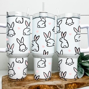 May include: White tumblers with a bunny and heart pattern. The tumblers have a white handle and lid, and a straw. The bunny design is black with pink ears and a white tail. The hearts are pink.