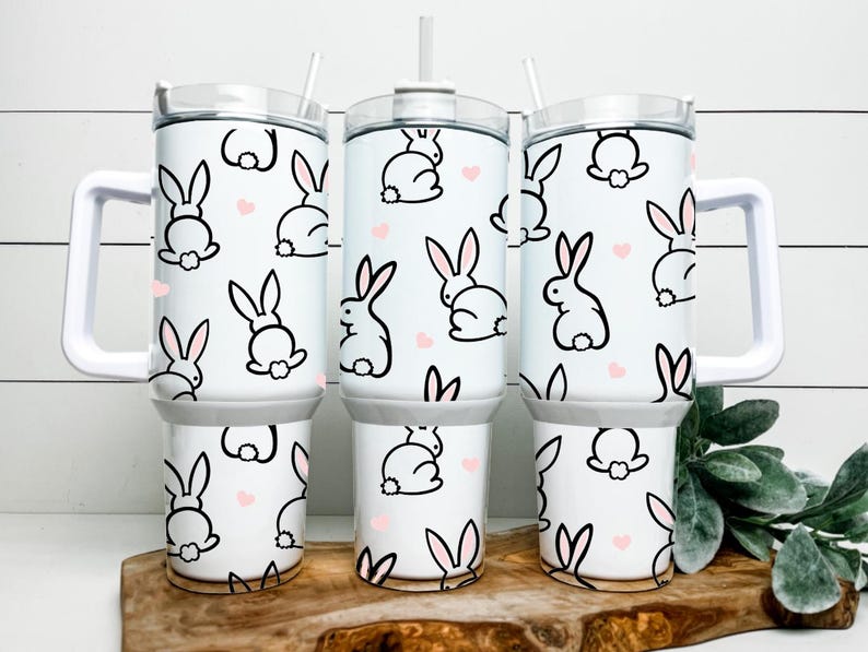 May include: White tumblers with a bunny and heart pattern. The tumblers have a white handle and lid, and a straw. The bunny design is black with pink ears and a white tail. The hearts are pink.