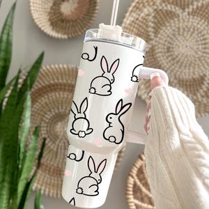 May include: A white tumbler with a handle, featuring a repeating pattern of black bunny illustrations and small pink hearts. The tumbler has a clear lid and a white straw. The background includes woven wall decor and a green plant.