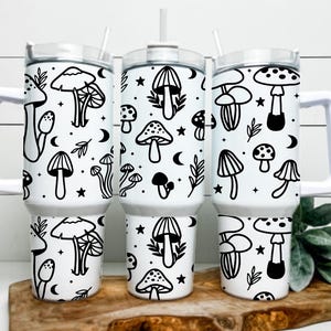 May include: Three white insulated tumblers with black mushroom and celestial designs. Each tumbler has a white handle, a clear lid, and a straw. The mushroom illustrations include various types, stars, and crescent moons.