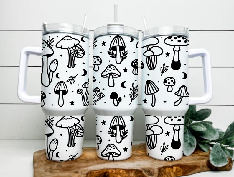 May include: Three white insulated tumblers with black mushroom and celestial designs. Each tumbler has a white handle, a clear lid, and a straw. The mushroom illustrations include various types, stars, and crescent moons.
