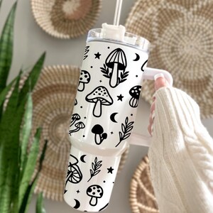 May include: A white travel tumbler with a black mushroom and celestial design. The tumbler features various mushroom illustrations, stars, and crescent moons. It has a white handle and a clear lid with a straw. The tumbler is designed for cold beverages.