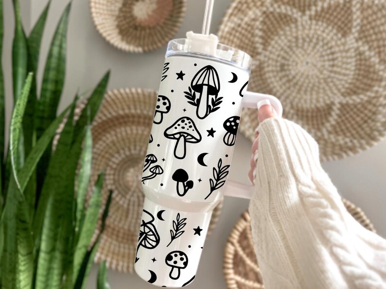 May include: A white travel tumbler with a black mushroom and celestial design. The tumbler features various mushroom illustrations, stars, and crescent moons. It has a white handle and a clear lid with a straw. The tumbler is designed for cold beverages.