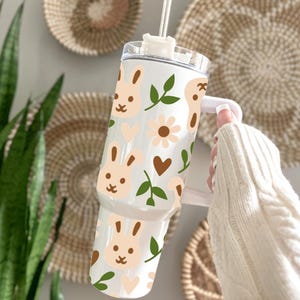 May include: A white tumbler with a bunny and floral pattern. The design includes light brown bunny faces, flowers, hearts, and green leaves. The tumbler has a white handle and a clear lid with a straw.