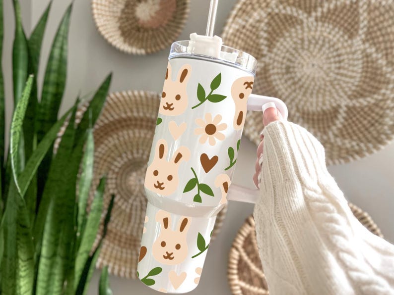 May include: A white tumbler with a bunny and floral pattern. The design includes light brown bunny faces, flowers, hearts, and green leaves. The tumbler has a white handle and a clear lid with a straw.