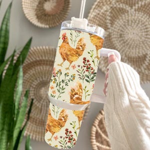 May include: A stainless steel tumbler with a chicken and floral pattern. The tumbler has a white handle and a clear lid with a straw. The design features watercolor illustrations of chickens, flowers, and foliage on a cream background.