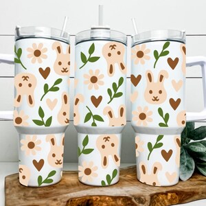 May include: Three white insulated tumblers with handles, decorated with a repeating pattern of light brown bunnies, flowers, and hearts, along with green leaves. Each tumbler has a white lid and a straw.