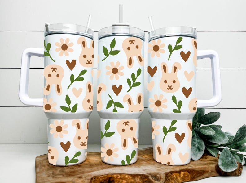 May include: Three white insulated tumblers with handles, decorated with a repeating pattern of light brown bunnies, flowers, and hearts, along with green leaves. Each tumbler has a white lid and a straw.