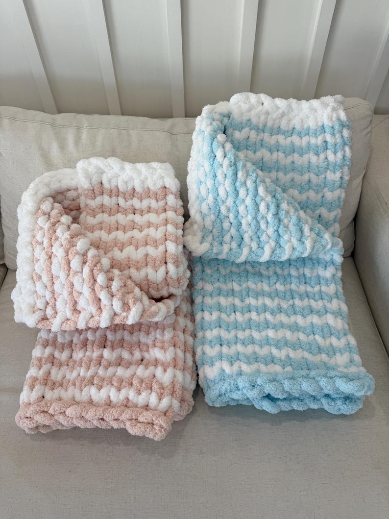 Peut inclure: Deux couvertures faites &agrave; la main, l'une rose et blanche, l'autre bleue et blanche. Les couvertures ont un motif boucl&eacute; textur&eacute;. Elles sont pli&eacute;es et pr&eacute;sent&eacute;es sur un canap&eacute; de couleur neutre.