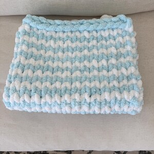 Peut inclure: Une couverture pour b&eacute;b&eacute; rectangulaire tricot&eacute;e &agrave; la main, dans un motif doux et textur&eacute;. La couverture pr&eacute;sente des rayures altern&eacute;es bleu clair et blanches, avec une bordure bleu clair uni. Design douillet pour une chambre d'enfant.