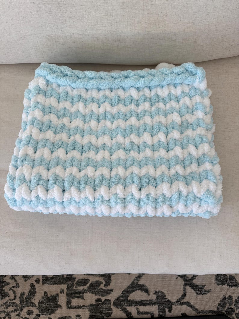 Peut inclure: Une couverture pour b&eacute;b&eacute; rectangulaire tricot&eacute;e &agrave; la main, dans un motif doux et textur&eacute;. La couverture pr&eacute;sente des rayures altern&eacute;es bleu clair et blanches, avec une bordure bleu clair uni. Design douillet pour une chambre d'enfant.