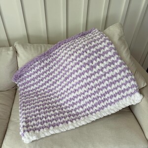 Peut inclure: Une couverture faite &agrave; la main avec un motif en chevrons. Elle pr&eacute;sente des rayures blanches et lavande altern&eacute;es, avec une bordure blanche. La couverture est pli&eacute;e sur un canap&eacute;.