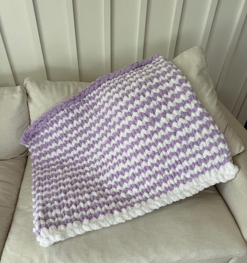 Peut inclure: Une couverture faite &agrave; la main avec un motif en chevrons. Elle pr&eacute;sente des rayures blanches et lavande altern&eacute;es, avec une bordure blanche. La couverture est pli&eacute;e sur un canap&eacute;.