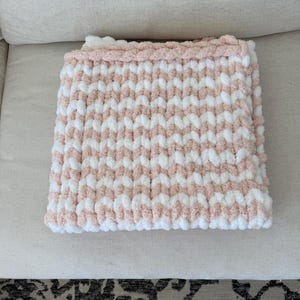 Peut inclure: Une couverture pli&eacute;e avec un motif ray&eacute; rose et blanc. La couverture est faite d'un fil &eacute;pais et textur&eacute;, avec une bordure rose unie. La couverture est soigneusement pli&eacute;e sur une surface de couleur claire.
