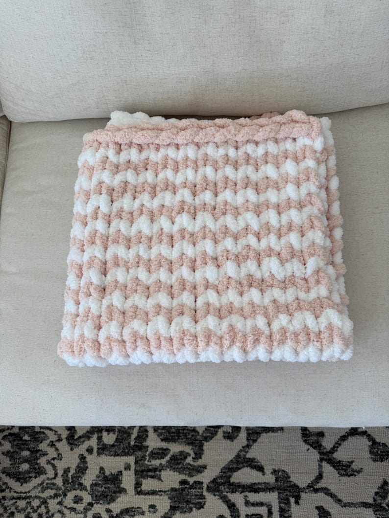 Peut inclure: Une couverture pli&eacute;e avec un motif ray&eacute; rose et blanc. La couverture est faite d'un fil &eacute;pais et textur&eacute;, avec une bordure rose unie. La couverture est soigneusement pli&eacute;e sur une surface de couleur claire.
