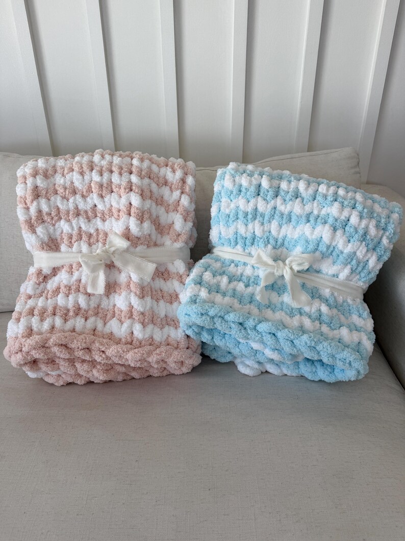 Peut inclure: Deux couvertures en tricot &eacute;pais, une rose et blanche, l'autre bleue et blanche. Chacune est attach&eacute;e avec un ruban blanc. Les couvertures sont pli&eacute;es et semblent &ecirc;tre faites d'une mati&egrave;re moelleuse, id&eacute;ale pour un b&eacute;b&eacute; ou un enfant.