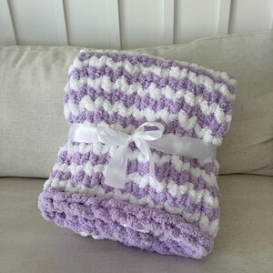 Peut inclure: Une couverture douce &agrave; motif chevron lavande et blanc, attach&eacute;e avec un ruban de satin blanc. La couverture est faite d'une mati&egrave;re moelleuse et textur&eacute;e, id&eacute;ale pour un b&eacute;b&eacute; ou comme jet&eacute; d&eacute;coratif. La couverture est pli&eacute;e et pr&ecirc;te &agrave; &ecirc;tre offerte.