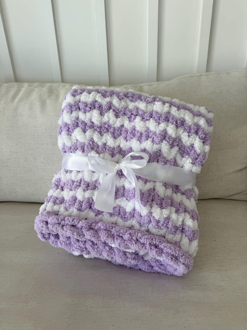 Peut inclure: Une couverture douce &agrave; motif chevron lavande et blanc, attach&eacute;e avec un ruban de satin blanc. La couverture est faite d'une mati&egrave;re moelleuse et textur&eacute;e, id&eacute;ale pour un b&eacute;b&eacute; ou comme jet&eacute; d&eacute;coratif. La couverture est pli&eacute;e et pr&ecirc;te &agrave; &ecirc;tre offerte.