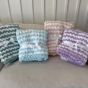 Peut inclure: Quatre couvertures en tricot &eacute;pais, vertes, bleues, roses et violettes, chacune avec des accents blancs. Les couvertures sont nou&eacute;es avec des rubans de satin blancs et sont pr&eacute;sent&eacute;es sur un canap&eacute; de couleur neutre.
