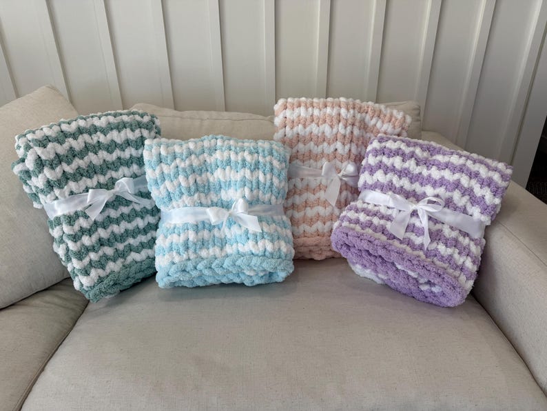 Peut inclure: Quatre couvertures en tricot &eacute;pais, vertes, bleues, roses et violettes, chacune avec des accents blancs. Les couvertures sont nou&eacute;es avec des rubans de satin blancs et sont pr&eacute;sent&eacute;es sur un canap&eacute; de couleur neutre.