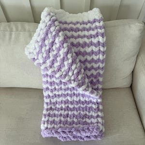 Peut inclure: Une couverture douce tricot&eacute;e &agrave; la main avec un motif en chevrons. La couverture pr&eacute;sente des rang&eacute;es altern&eacute;es de fil blanc et lavande. La couverture est pli&eacute;e sur un canap&eacute; de couleur claire, mettant en valeur sa texture et son design.