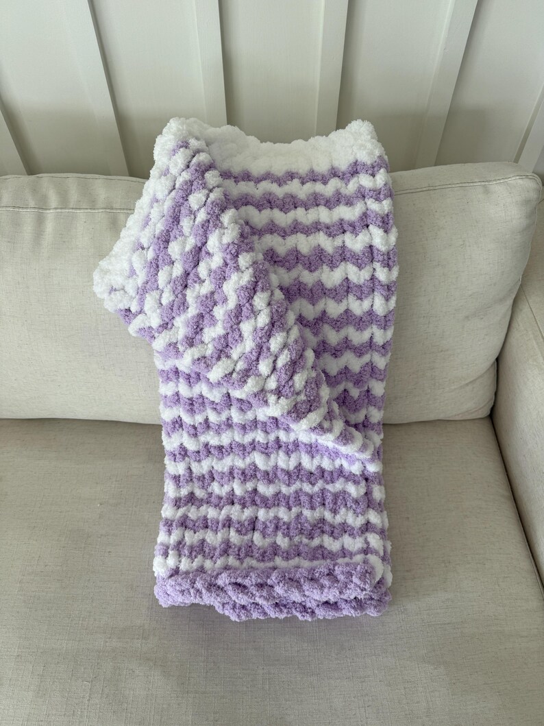 Peut inclure: Une couverture douce tricot&eacute;e &agrave; la main avec un motif en chevrons. La couverture pr&eacute;sente des rang&eacute;es altern&eacute;es de fil blanc et lavande. La couverture est pli&eacute;e sur un canap&eacute; de couleur claire, mettant en valeur sa texture et son design.