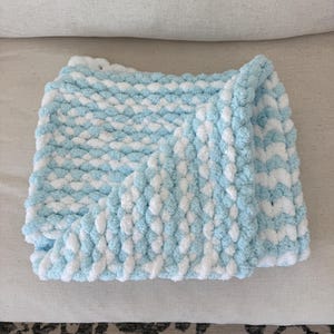 Peut inclure: Une couverture pour b&eacute;b&eacute; tricot&eacute;e &agrave; la main, &agrave; rayures bleues et blanches. Fabriqu&eacute;e avec une laine douce et textur&eacute;e, elle est pli&eacute;e pour montrer la texture et les variations de couleurs. Id&eacute;ale pour un cadeau.