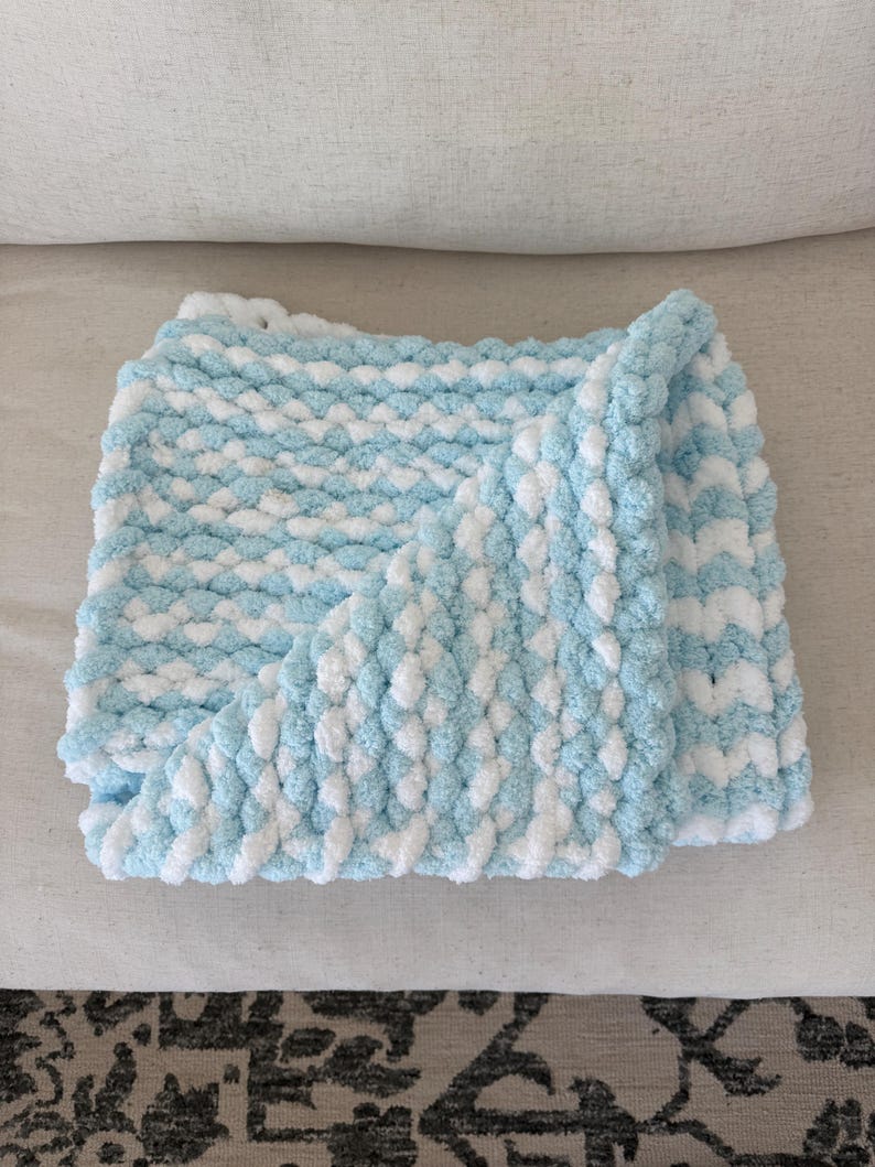 Peut inclure: Une couverture pour b&eacute;b&eacute; tricot&eacute;e &agrave; la main, &agrave; rayures bleues et blanches. Fabriqu&eacute;e avec une laine douce et textur&eacute;e, elle est pli&eacute;e pour montrer la texture et les variations de couleurs. Id&eacute;ale pour un cadeau.