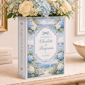 May include: A rectangular book-shaped vase featuring a coastal scene with blue hydrangeas, sailboats, and the text "The Wedding of Charlotte & Benjamin 05.18.2025". The vase is filled with a floral arrangement of white roses and blue flowers.