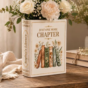 May include: A white ceramic vase shaped like a book, with the text "Just One More Chapter" and "Sophia's Library". The vase is filled with white roses, pink peonies, and greenery. The vase is sitting on a wooden surface.