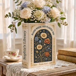 Könnte beinhalten: Eine cremefarbene Keramikvase in Buchform, gefüllt mit weißen Rosen, blauen Hortensien und Grünzeug. Die Vase hat ein Blumenmuster und den Text "Bloom Where God Planted You" und "Sophia's Library". Eine Tasse und Untertasse sind ebenfalls zu sehen.