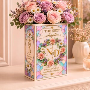 Pode incluir: Um arranjo floral de rosas cor-de-rosa e roxas, margaridas e vegetação em cima de uma caixa decorativa. A caixa apresenta um design de vitral com o texto "THE NEXT chapter" e "N&D". A caixa também tem o texto "Forever and always, together for a lifetime".