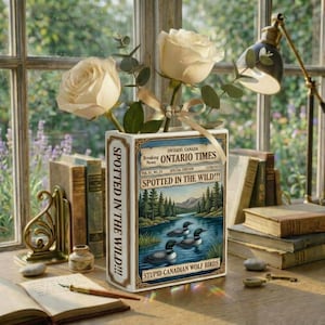 May include: A decorative vase shaped like a book, featuring the text "Spotted in the Wild!!!" and "Ontario Times." The vase holds white roses and greenery, with a ribbon and butterfly accent. The image includes other books, a desk lamp, and a window.