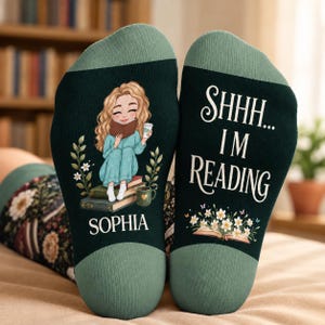 May include: A pair of dark green socks with light green cuffs. The left sock features a cartoon of a blonde person reading a book while sitting on a stack of books, with the name SOPHIA printed below. The right sock displays the text SHHH... I'M READING above an open book with white flowers.