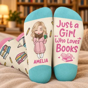 May include: A pair of cream-colored socks with teal cuffs. One sock features a cartoon of a blonde person reading a book with the name AMELIA printed below. The other sock displays the text Just a Girl Who Loves Books with a small book icon.