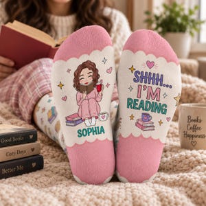 May include: A pair of white and pink socks featuring a cartoon illustration of a person reading a book with the name Sophia printed below. The other sock displays the text SHHH... I'M READING along with illustrations of books, a mug, and stars.