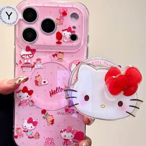 Puede incluir: Funda de teléfono rosa con una superposición transparente. Presenta un patrón repetido de personajes de Hello Kitty, corazones y otros elementos decorativos. Un dije de Hello Kitty transparente separado está adjunto.