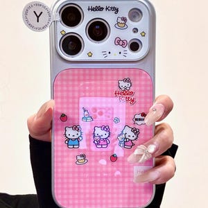 May include: A pink and silver Hello Kitty phone case. The case has a pink gingham pattern and various Hello Kitty illustrations. The top section displays the Hello Kitty logo and camera apertures. The case is designed to protect a mobile phone.