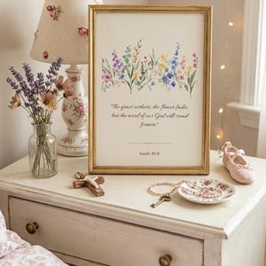 May include: A framed print with a gold frame displays a watercolour floral design in purple, blue, and yellow. The text reads, "The grass withers, the flower fades, but the word of our God will stand forever." A lamp, flowers, and decorative items are on a white bedside table.