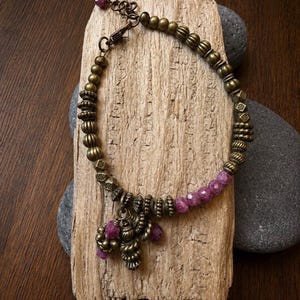 May include: A bracelet featuring antique brass-toned metal beads with textured surfaces and faceted, deep pinkish-purple gemstone beads. The design includes a central cluster of dangling beads and a metal lobster claw clasp with an adjustable chain.