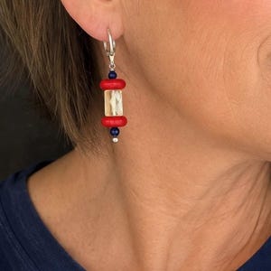 May include: A drop earring featuring a silver hoop at the top, followed by a small dark blue bead, a bright red disc bead, a central clear faceted rectangular bead, another red disc bead, and a final small dark blue bead with a silver accent.