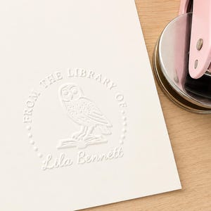 May include: A light pink metal embosser tool positioned next to a white paper sheet featuring a circular embossed design. The design includes an owl perched on a book, surrounded by the text FROM THE LIBRARY OF and the name Lila Bennett.