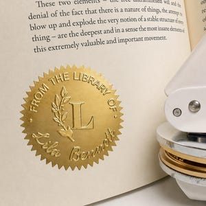 May include: A gold circular embossed seal with a serrated edge pressed onto a book page. The seal features the text FROM THE LIBRARY OF and Lila Bennett, with a central letter L and a leafy branch design, next to a white metal embosser tool.
