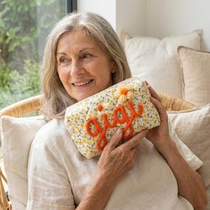 May include: A floral print clutch with the name "Gigi" in orange script. The clutch features a yellow and white floral pattern with a small orange flower. The clutch is rectangular and appears to be made of fabric.
