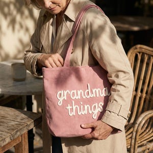 May include: A dusty rose corduroy tote bag featuring the white embroidered text "grandma things" on the front. The bag has two shoulder straps and a rectangular shape, constructed from a textured fabric with vertical ribbed lines throughout.