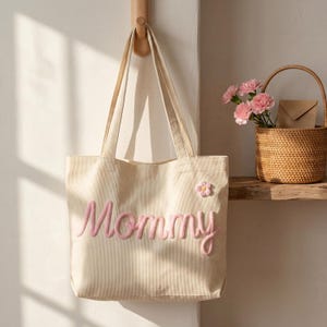 May include: A cream-colored corduroy tote bag featuring the word Mommy embroidered in pink script lettering across the front. A small, pink and white fabric flower applique is attached to the upper right side of the bag, which has two handles.