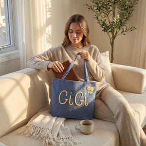 May include: A blue denim tote bag featuring the name GiGi embroidered in yellow script lettering on the front, accompanied by a small yellow and white flower applique. The bag has two matching blue fabric handles and a rectangular shape.